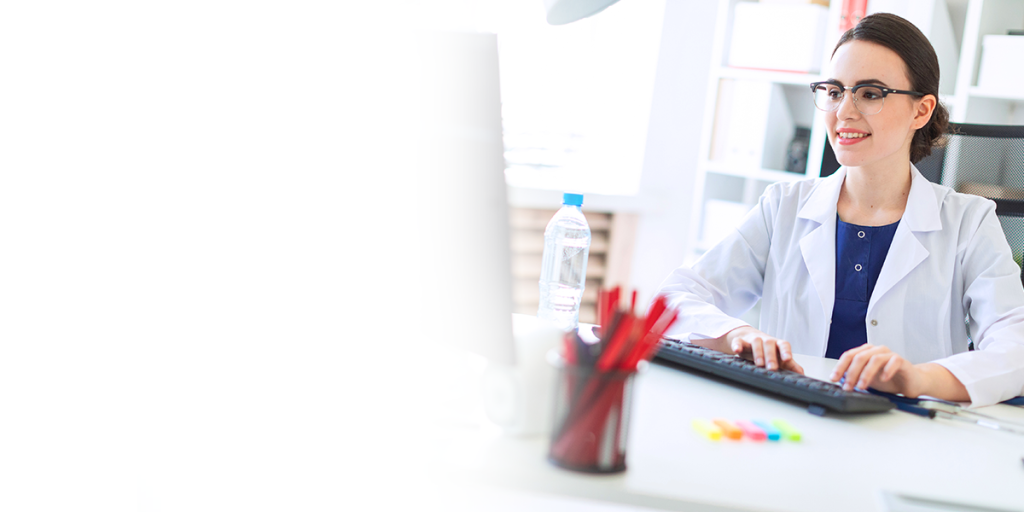 A female dentist smiling while using a computer. She is sitting at a desk, wearing a lab coat and glasses.